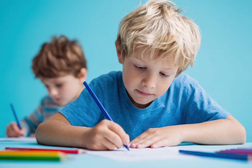 Young boy is drawing with blue pencil on white paper, while another child is engaged in creative activity in the background, highlighting a vibrant learning environment